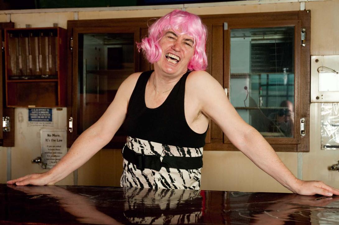 A figure at a bar wearing a bright pink wig with a wide smile.